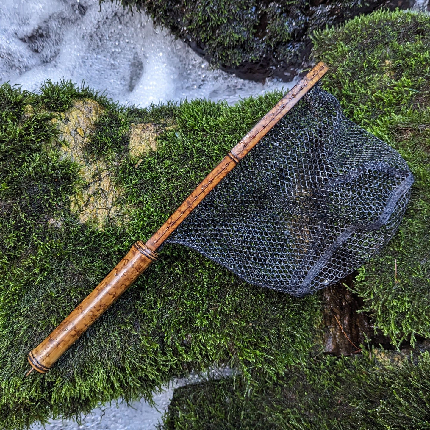 A bamboo fly fishing net lying on a rock in the water on the river in Western North Carolina. The rubber net basket is smooth and shiny and protects the fish for catch and release. The handle is flame cured for strength and durability and there is copper stitching and a copper keeper ring to attach your net keeper to.