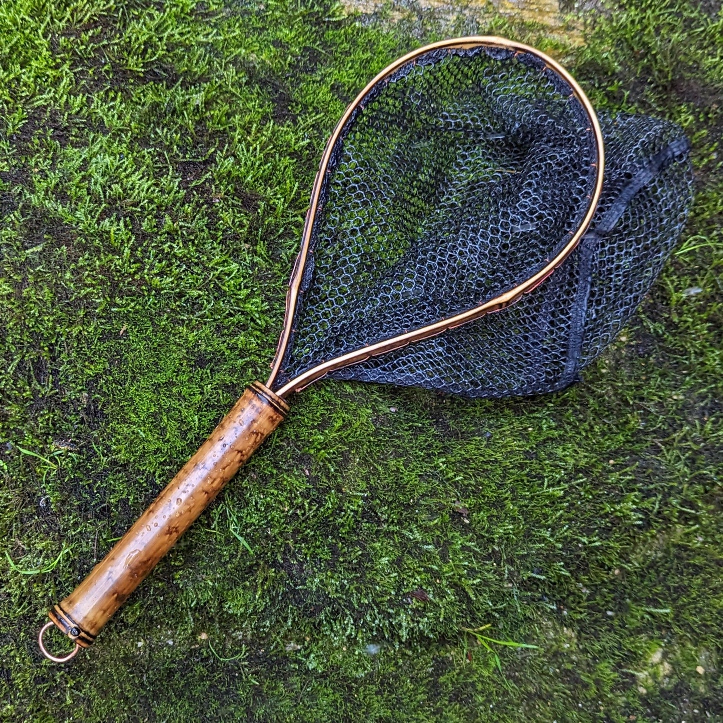 A bamboo fly fishing net lying on a rock in the water on the river in Western North Carolina. The rubber net basket is smooth and shiny and protects the fish for catch and release. The handle is flame cured for strength and durability and there is copper stitching and a copper keeper ring to attach your net keeper to.