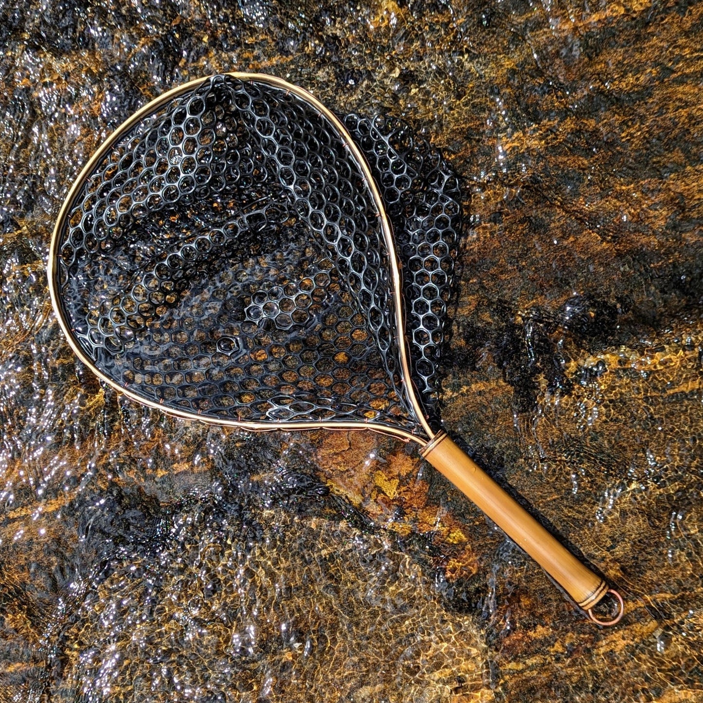 A bamboo fly fishing net lying on a rock in the water on the river in Western North Carolina. The rubber net basket is smooth and shiny and protects the fish for catch and release. The handle is flame cured for strength and durability and there is copper stitching and a copper keeper ring to attach your net keeper to.