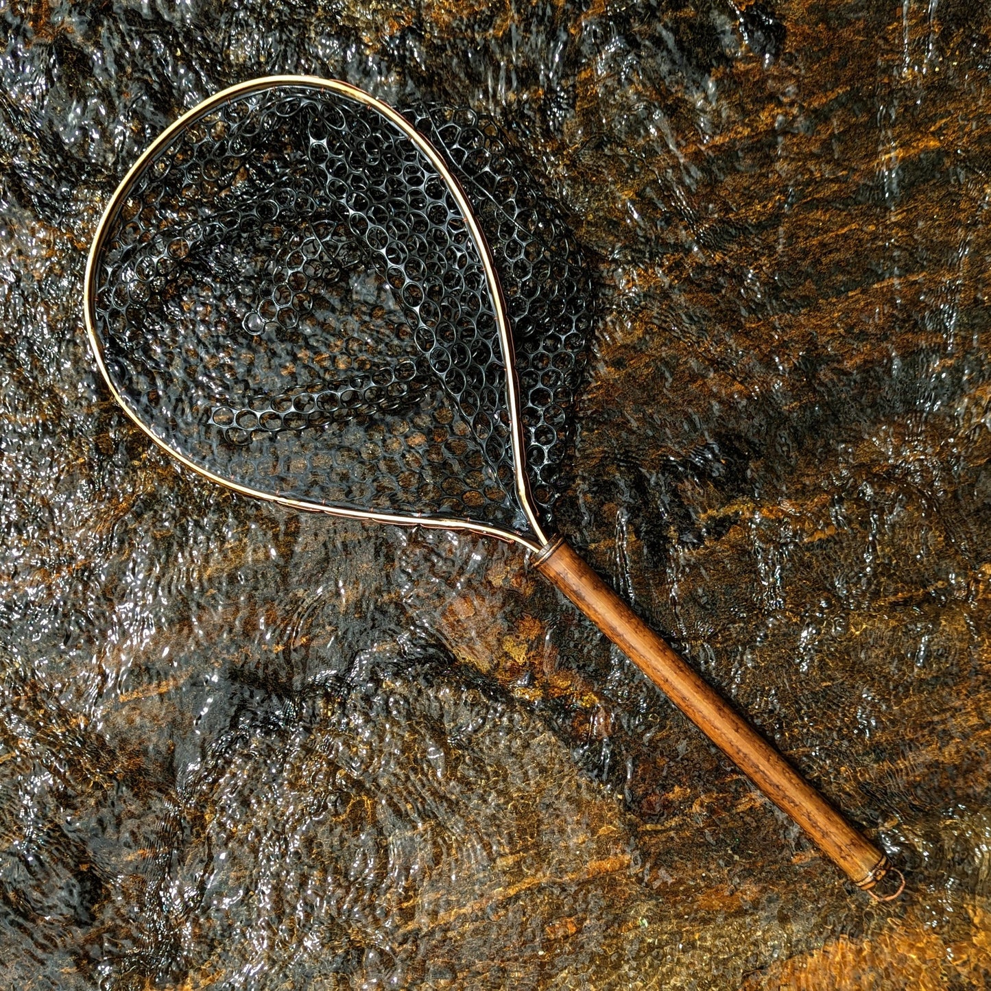 A bamboo fly fishing net lying on a rock in the water on the river in Western North Carolina. The rubber net basket is smooth and shiny and protects the fish for catch and release. The handle is flame cured for strength and durability and there is copper stitching and a copper keeper ring to attach your net keeper to.