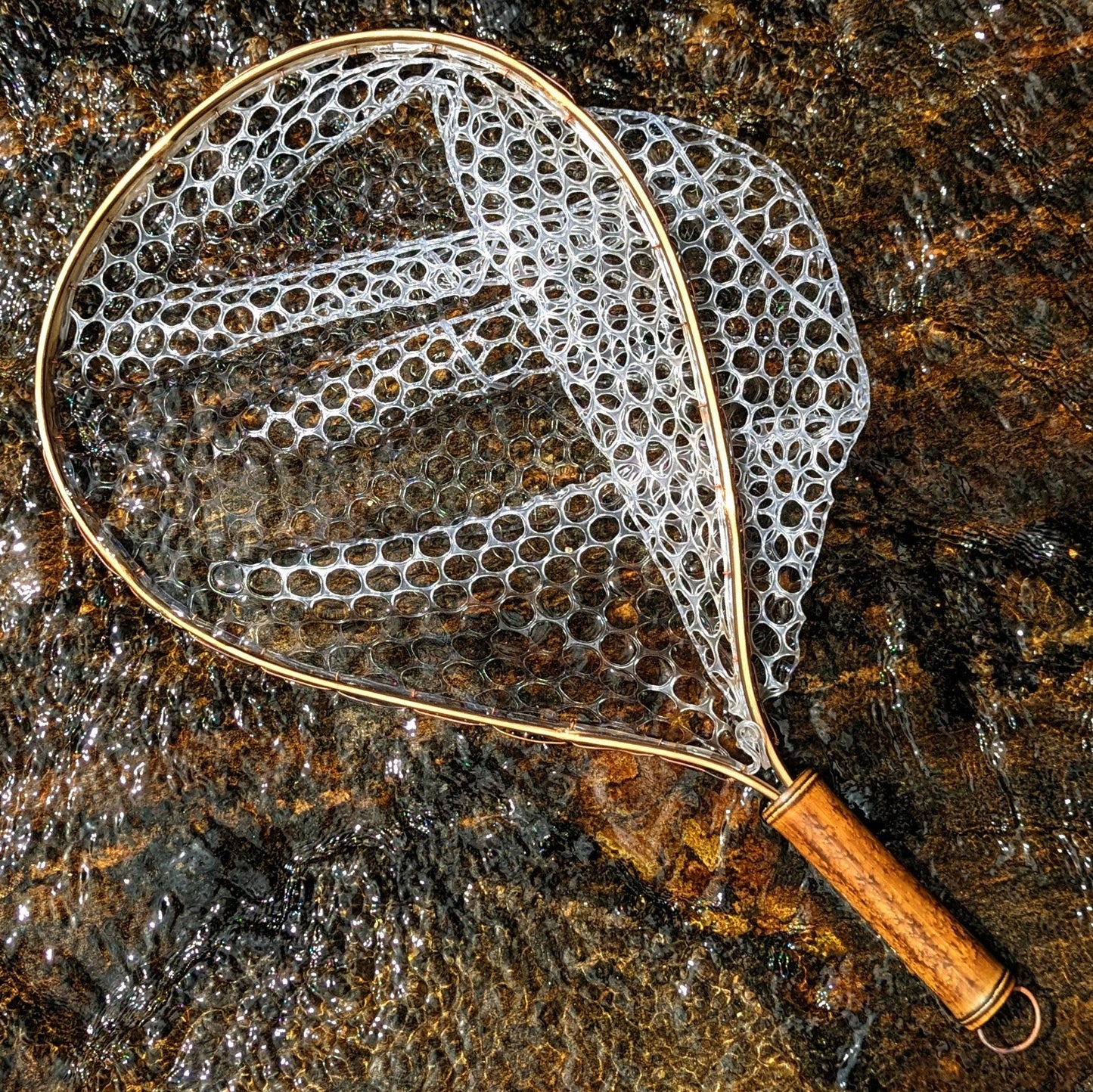 A bamboo fly fishing net lying on a rock in the water on the river in Western North Carolina. The rubber net basket is smooth and shiny and protects the fish for catch and release. The handle is flame cured for strength and durability and there is copper stitching and a copper keeper ring to attach your net keeper to.