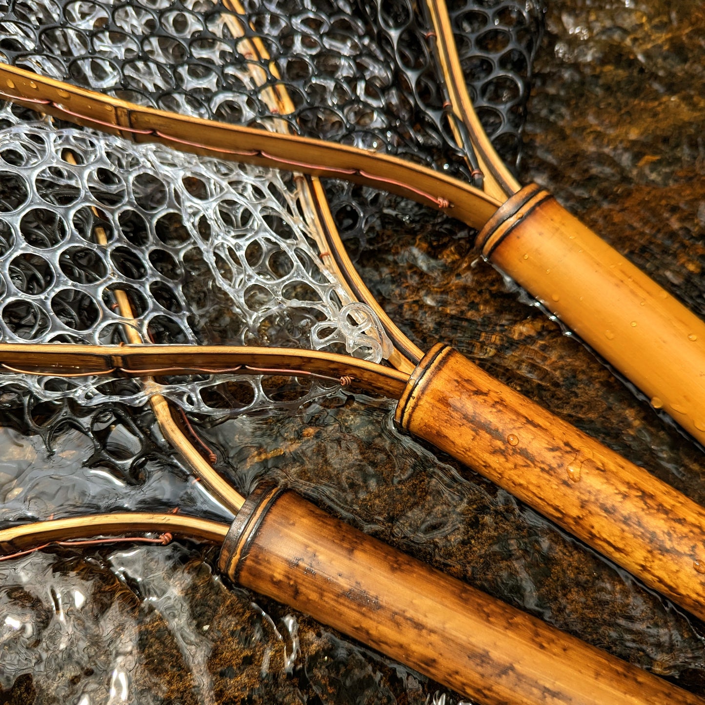 A bamboo fly fishing net lying on a rock in the water on the river in Western North Carolina. The rubber net basket is smooth and shiny and protects the fish for catch and release. The handle is flame cured for strength and durability and there is copper stitching and a copper keeper ring to attach your net keeper to.