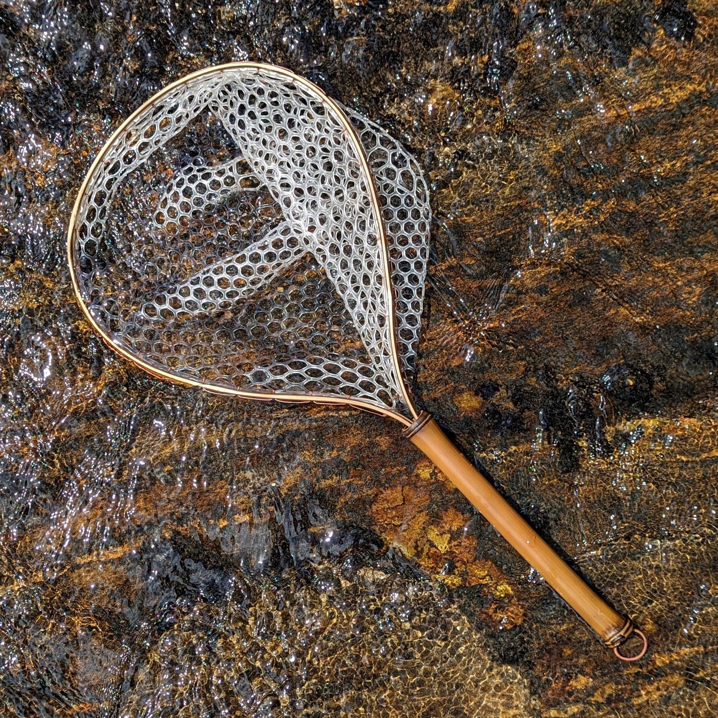 A bamboo fly fishing net lying on a rock in the water on the river in Western North Carolina. The rubber net basket is smooth and shiny and protects the fish for catch and release. The handle is flame cured for strength and durability and there is copper stitching and a copper keeper ring to attach your net keeper to.