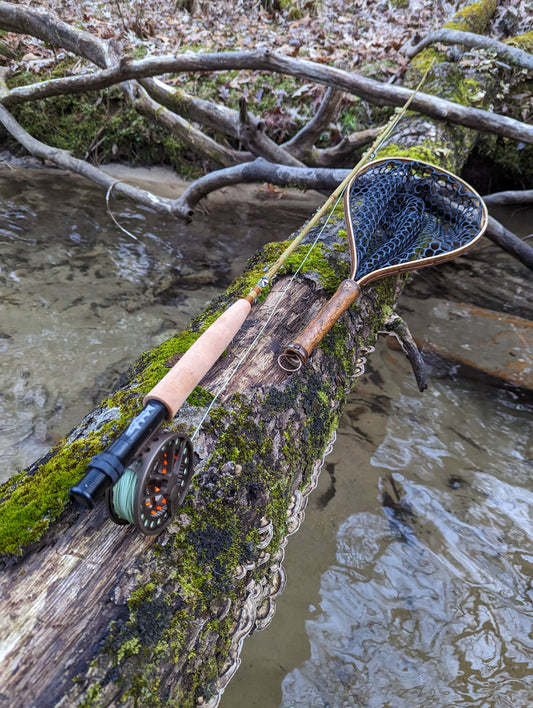 A bamboo fly fishing net on a log over the river next to a fly fishing pole