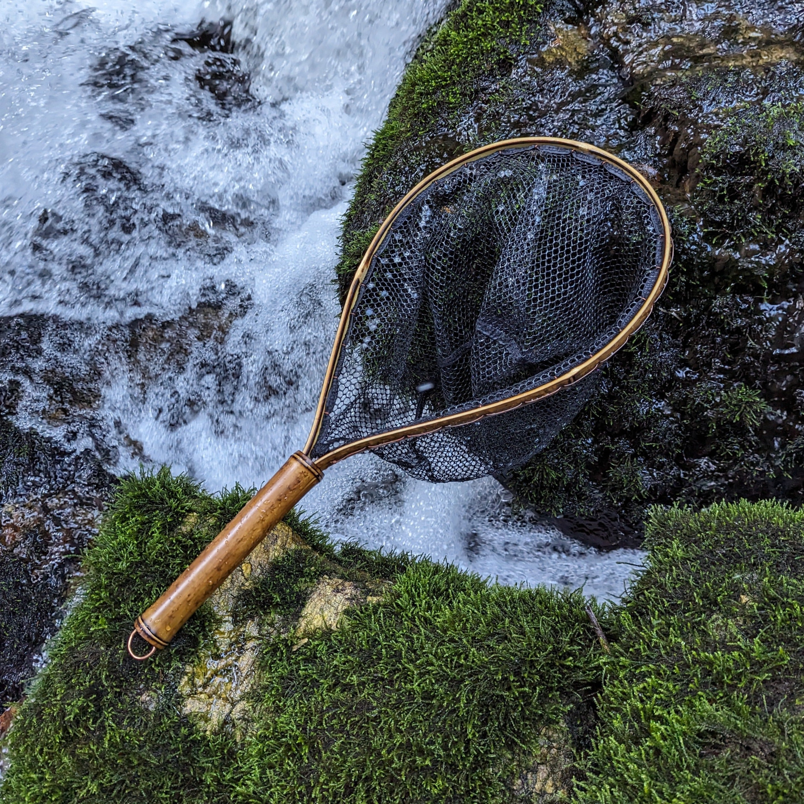 A bamboo fly fishing net lying on a rock in the water on the river in Western North Carolina. The rubber net basket is smooth and shiny and protects the fish for catch and release. The handle is flame cured for strength and durability and there is copper stitching and a copper keeper ring to attach your net keeper to.