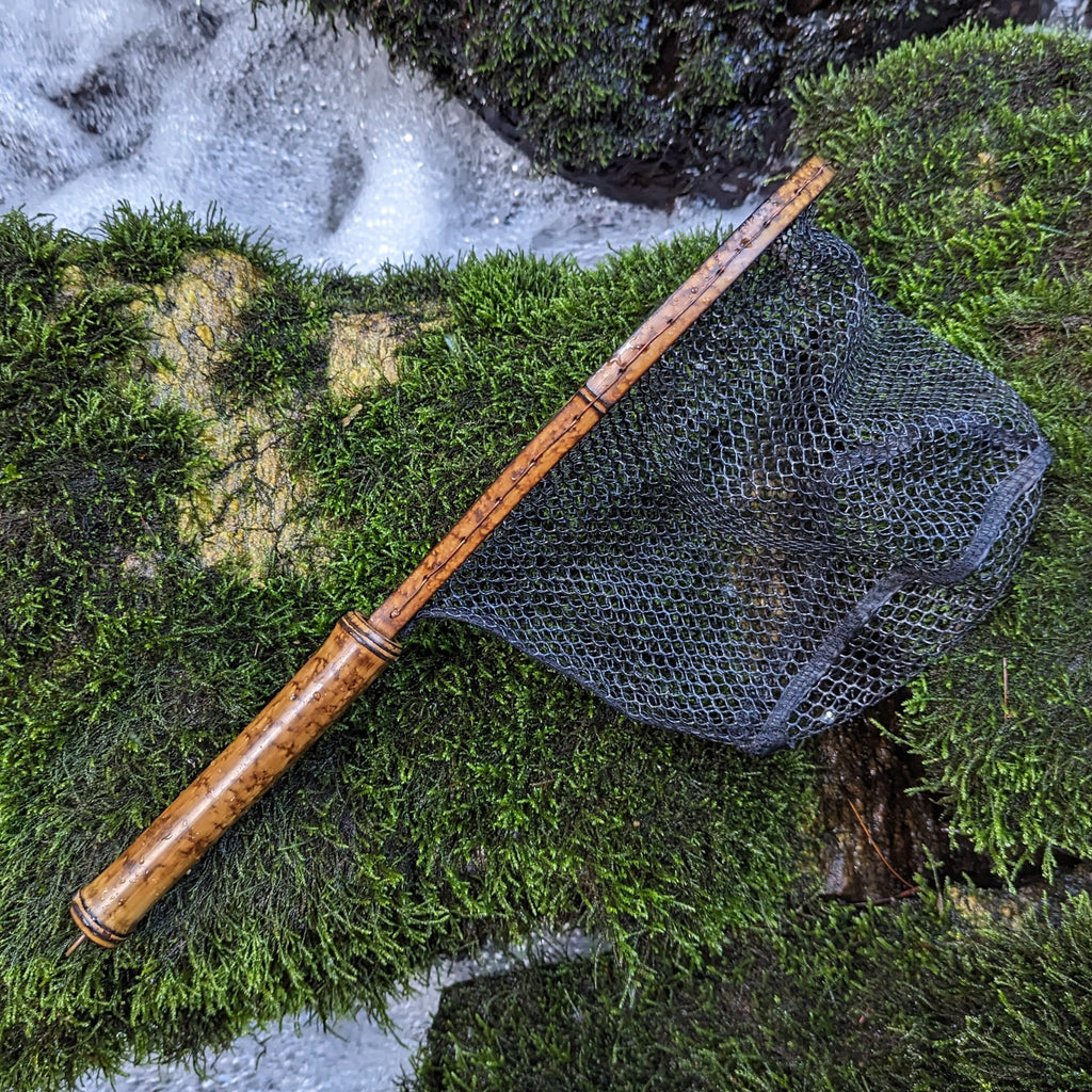 A bamboo fly fishing net lying on a rock in the water on the river in Western North Carolina. The rubber net basket is smooth and shiny and protects the fish for catch and release. The handle is flame cured for strength and durability and there is copper stitching and a copper keeper ring to attach your net keeper to.