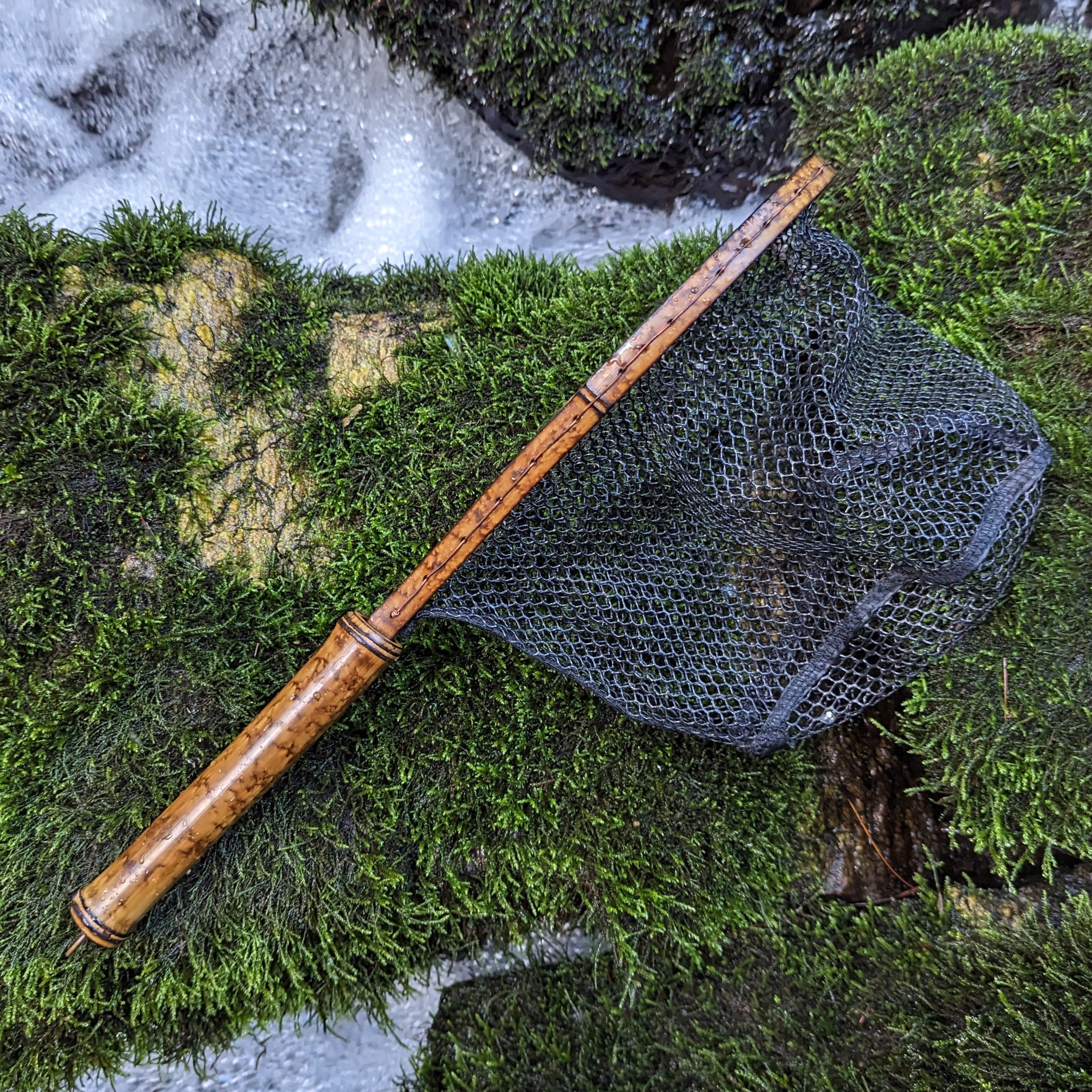 A bamboo fly fishing net lying on a rock in the water on the river in Western North Carolina. The rubber net basket is smooth and shiny and protects the fish for catch and release. The handle is flame cured for strength and durability and there is copper stitching and a copper keeper ring to attach your net keeper to.