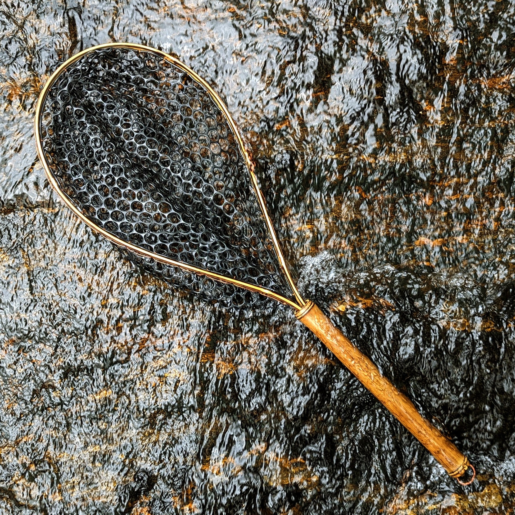 A bamboo fly fishing net lying on a rock in the water on the river in Western North Carolina. The rubber net basket is smooth and shiny and protects the fish for catch and release. The handle is flame cured for strength and durability and there is copper stitching and a copper keeper ring to attach your net keeper to.