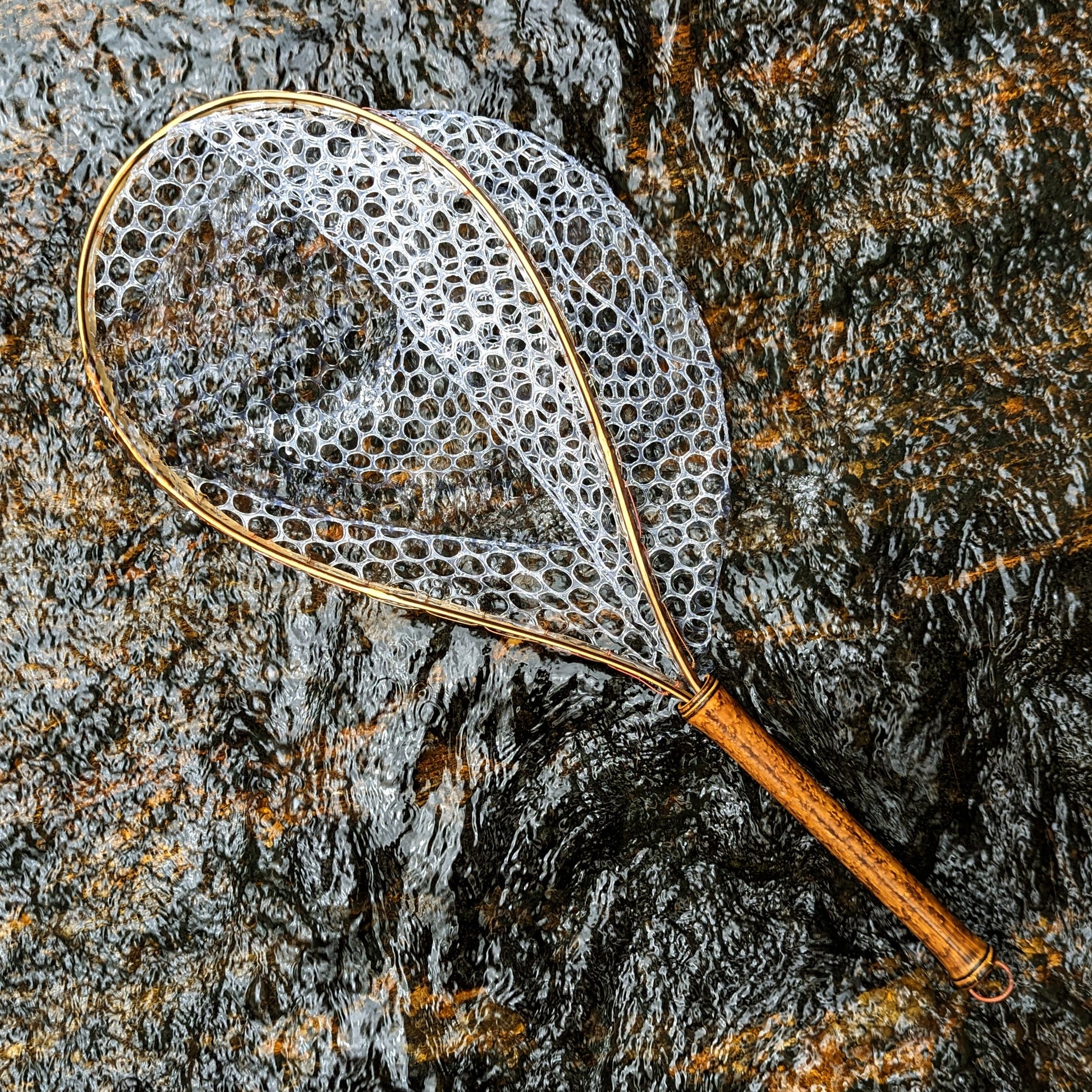 A bamboo fly fishing net lying on a rock in the water on the river in Western North Carolina. The rubber net basket is smooth and shiny and protects the fish for catch and release. The handle is flame cured for strength and durability and there is copper stitching and a copper keeper ring to attach your net keeper to.