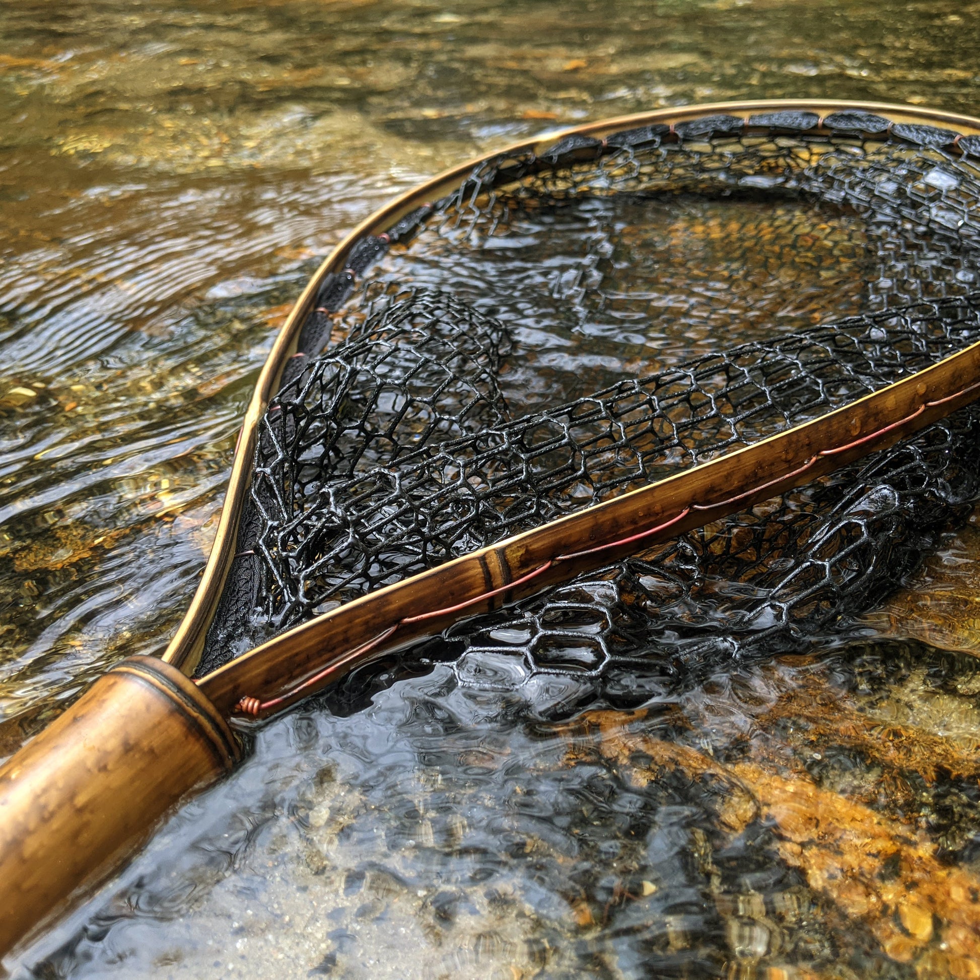 A bamboo fly fishing net lying on a rock in the water on the river in Western North Carolina. The rubber net basket is smooth and shiny and protects the fish for catch and release. The handle is flame cured for strength and durability and there is copper stitching and a copper keeper ring to attach your net keeper to.