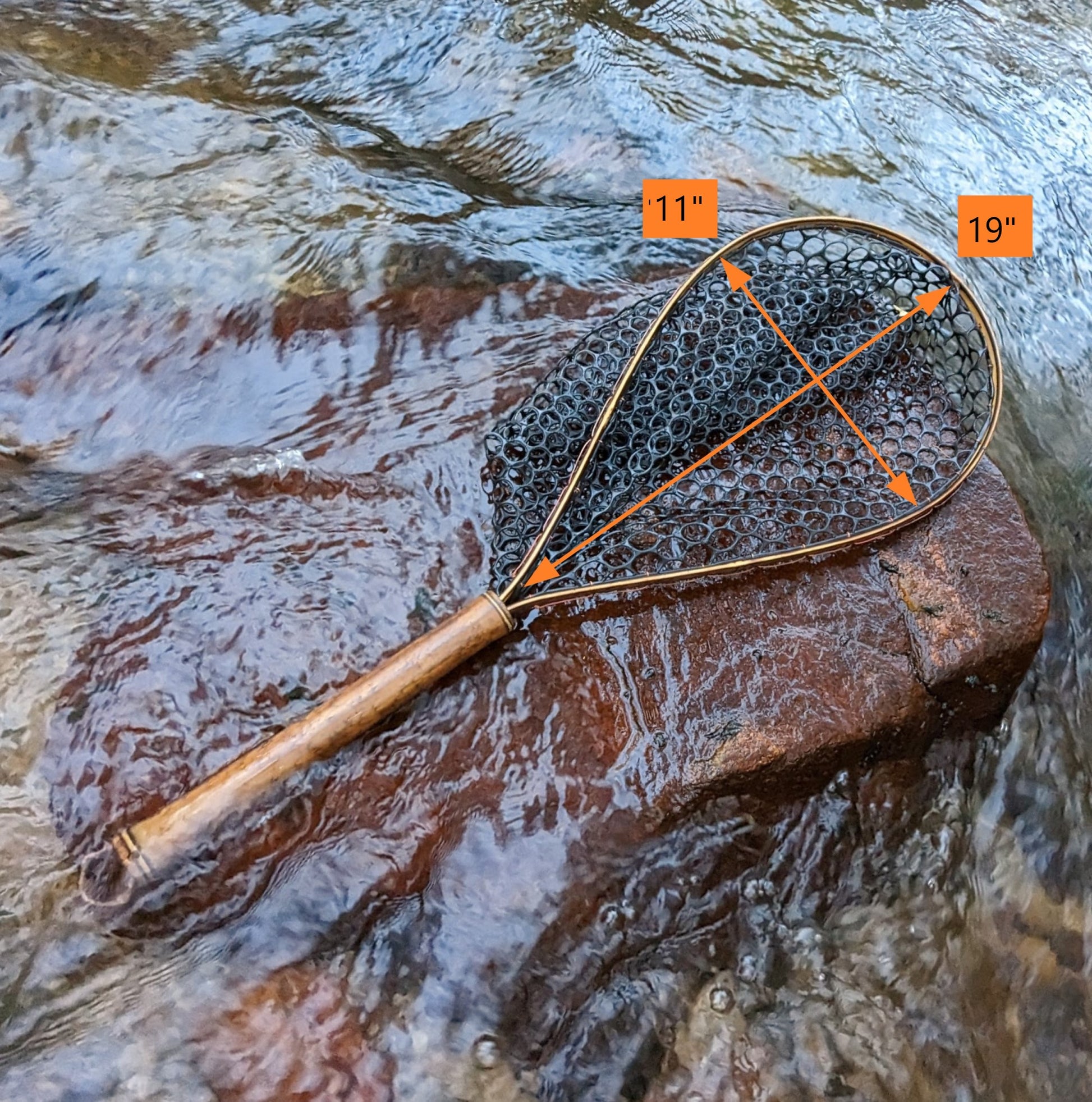 A bamboo fly fishing net lying on a rock in the water on the river in Western North Carolina. The rubber net basket is smooth and shiny and protects the fish for catch and release. The handle is flame cured for strength and durability and there is copper stitching and a copper keeper ring to attach your net keeper to.