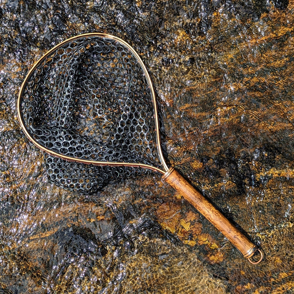A bamboo fly fishing net lying on a rock in the water on the river in Western North Carolina. The rubber net basket is smooth and shiny and protects the fish for catch and release. The handle is flame cured for strength and durability and there is copper stitching and a copper keeper ring to attach your net keeper to.